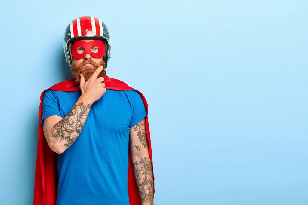 People and super power concept. Serious man with red thick beard, wears helmet and red superhero cap